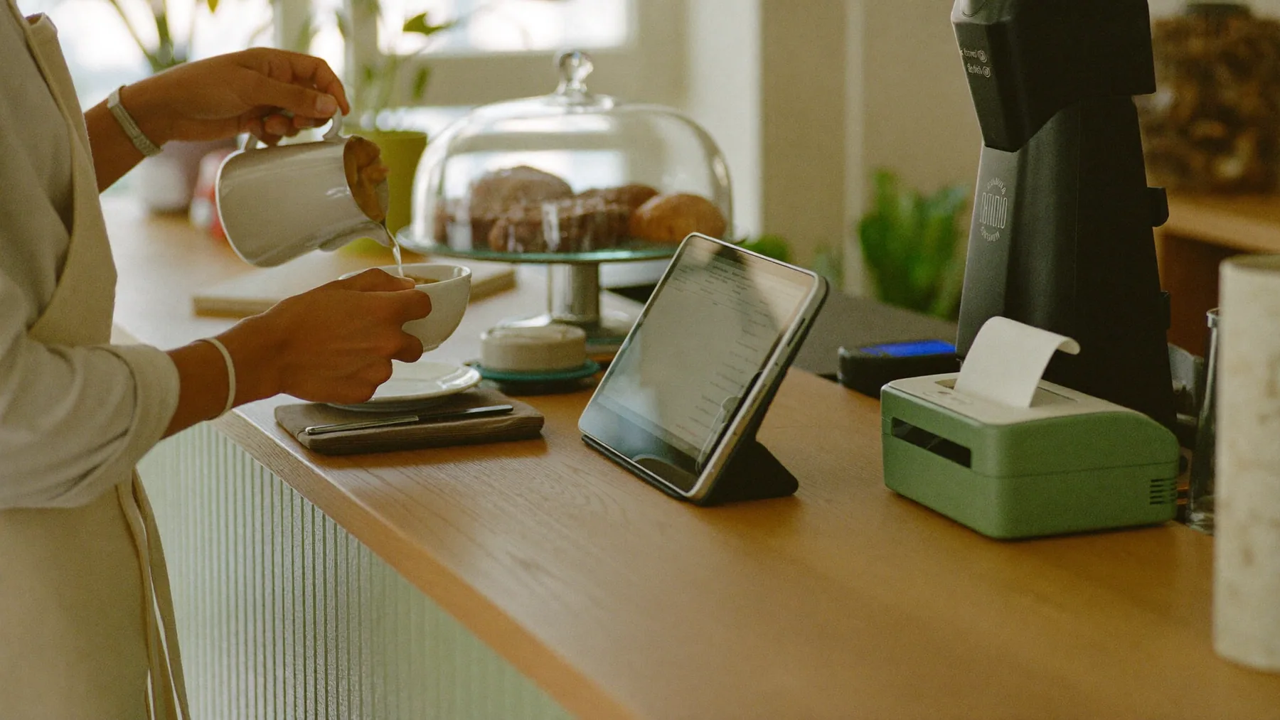 Barista sirviendo un café en el mostrador de una cafetería, con la tablet de cobro y la impresora al lado