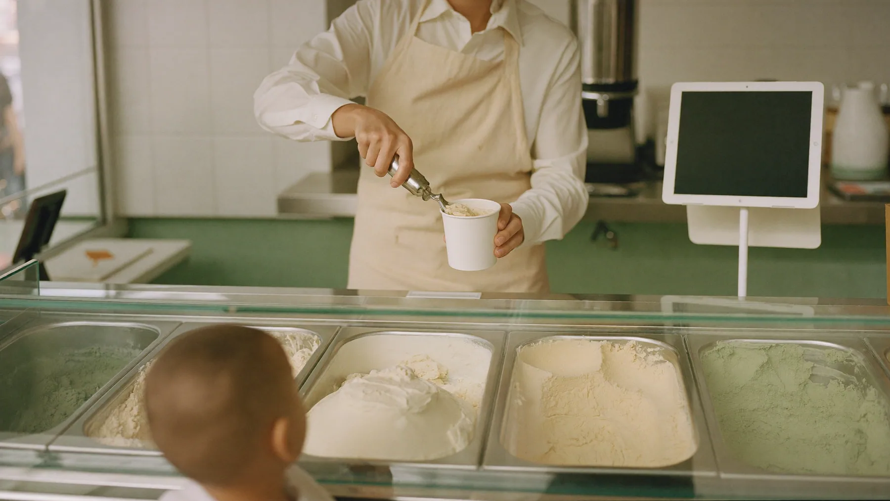 Heladero sirviendo un pote frente a la vitrina de gustos, con la tablet de cobro al fondo