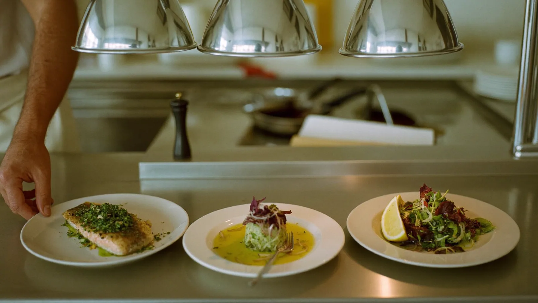 Tres platos listos bajo las lámparas del pase de una cocina profesional
